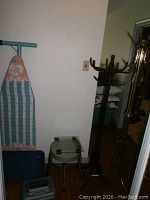 Photo showing ironing board with crocheted cover, vintage doctor's stool, glass-top scale, and two wooden coat trees.