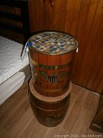 Two wooden Americana style storage barrels stacked in corner of room, top barrel decorated with eagle and shield, bottom barrel solid wood with removable lids.