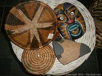 Large white wicker basket with lid holding trays and collectibles on top including a tribal mask and fabric wood decorative item.