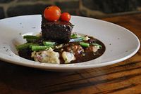 Braised beef entree plated with mashed potatoes, green beans, cherry tomatoes, and sauce on a white plate against a stone wall background.