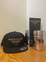 Photo of black MacKinnon Brothers Brewing baseball cap with yellow and white embroidery, accompanied by two stainless steel barrel-shaped drinking mugs with MacKinnon Brothers Brewing and Back Farm branding, on a wooden surface.