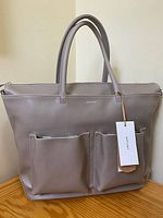 Front view of light grey Matt & Nat diaper bag with two front pockets and hang tag showing brand name.