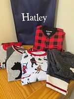 Photo of 6 pieces of Hatley children's pajamas size 6 laid on table, showing variety of patterns including buffalo plaid, wildlife winter scenes, and bear silhouette with Hatley bag in background.