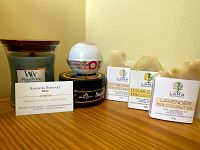 Photo showing the body care items on a wooden surface: WoodWick candle, bath bomb, Bee By the Sea sugar scrub tin, and three L.A.M.A soaps, each labeled with scent and coconut oil content.