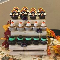 Display of various decorated cupcakes including chocolate mint, pumpkin spice, and chocolate nut with turkey toppers arranged on a tiered white stand decorated with orange and red fall leaves.