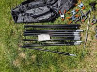 Photo showing several black metal poles, clamps, and a carrying bag, likely parts of tripod, risers, light stands, or background support equipment.