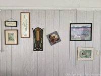 Wall with assorted framed art pieces including botanical prints, horseshoe plaque, butterfly tile, and landscape paintings, arranged for decorative display.