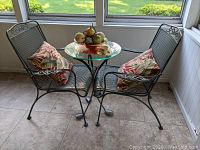 Photo of small patio table with glass top and two metal mesh chairs with floral cushions, viewed from side angle