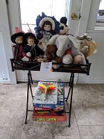 Small black metal folding table with dolls, stuffed animals, and boxed games displayed on top and underneath.