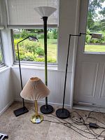 Photo shows three floor lamps and one table lamp grouped on a tile floor near windows. Two black adjustable floor lamps with rectangular bases, one tall green and black floor lamp with a white conical shade, and one brass table lamp with beige pleated shade.