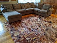 Front view of the carpet rug laid flat on a wooden floor under a sectional sofa (not included). The carpet is primarily brown with geometric patterns including triangles and swirls.