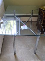 Rectangular glass top table with chrome metal legs and crossbar support on beige carpet next to a wall and a chair.