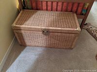 Photo of a rectangular wicker storage chest placed on carpet, showing the front and top with diamond weave pattern, metal latch, and brass corner guards.