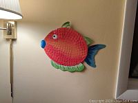 Orange textured metal fish wall decoration with green fins mounted on wall beside a lamp