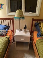 Night stand placed between two beds with green and orange bedding, topped with a standing lamp having a woven shade with blue fringe and a small sneaker used as decoration.
