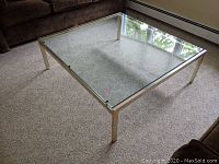 Square glass coffee table showing top glass surface and metal frame in room with carpet and couch background.