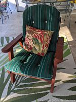 Front view of the redwood wooden outdoor chair with green striped cushions and floral decorative pillow on a green patterned rug.