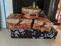 Front view of a stack of six decorative storage boxes with floral patterns and lids, including black fabric boxes with white flowers and rose patterned boxes with lids.