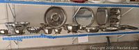 Shelf with 12 metal serving bowls, trays, and dishes in various sizes and shapes, some with footed bases and scalloped edges.