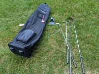 Photo showing black golf travel bag with wheels and pockets alongside four golf clubs and a small plastic putter returner device on grass