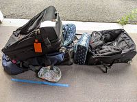 Photo showing large and small luggage and travel bags stacked, including a large black wheeled suitcase and smaller bags with patterns.