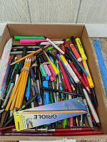 Cardboard box filled with a variety of pens, pencils, markers, and a pack of Dixon Oriole pencils among them.