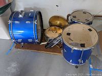 Blue beginner drum kit with bass drum, snare drum, floor tom, and two cymbals several with steel hardware arranged on a wooden platform.