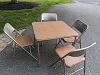Full set of square folding card table and four folding chairs placed outdoors on pavement near grass and stone pavers.