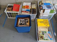 Five large plastic bins filled with assorted nonfiction and fiction books in hard and soft covers, stacked in a warehouse setting.