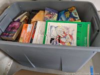 A grey plastic bin filled with multiple hardcover and softcover books showing a visible variety of titles and subjects.