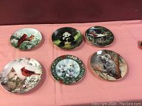 Overview of six assorted collector plates arranged on pink cloth, featuring birds, panda, and koala subjects in nature settings