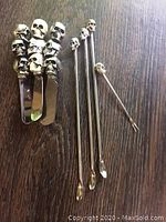 Photo showing eight metal serving utensils on a wooden surface. Four spreaders with flat blades topped with skulls, three thin stir spoons with small oval bowls and skull handles, and one two-pronged pickle fork with a skull handle.