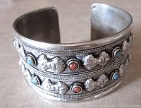 Front view showing the detailed embossed designs and alternating small turquoise and coral coloured cabochons on the wide silver tone cuff bangle.