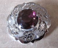 Front view of round silver tone Mizpah brooch with engraved acorns and leaves, large faceted purple glass stone center, approximately 1.5 inches across.
