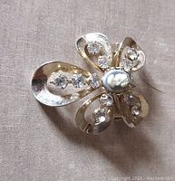 Front view showing flower-shaped brooch with five petals each adorned with clear rhinestones and central white cameo on blue-colored background.