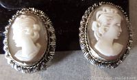 Close-up of the two oval shell cameo clip earrings showing carved female profiles framed by a textured silver/brass-tone metal border.