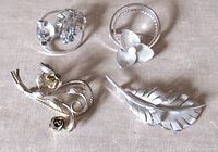 Four Bond Boyd sterling silver brooches arranged facing upward on a neutral fabric background, visible front details of each brooch showcasing stones, pearl, matte finish, and gilded roses.