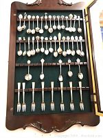 Frontal photo of wooden cabinet showing rows of collectible spoons and pickle forks mounted on green felt backing inside glass door.