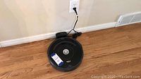 Top view of black Roomba vacuum on hardwood floor next to charging dock plugged into wall outlet