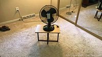Photo of small wooden table with black oscillating fan placed on it in room with carpet and mirrored closet doors.