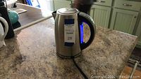 Stainless steel electric tea kettle on a kitchen countertop with a visible cord and blue light indicator shape visible in the handle.