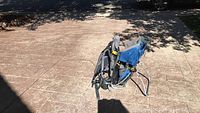 Side and angled view of the blue and gray Deuteronomy kid comfort backpack carrier standing on paved ground under partial shade, showing overall shape, stand, and straps.