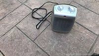 Small grey and black electric space heater placed on stone tile floor with visible top control dials and power cord.