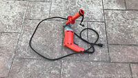Full view of the red Milwaukee drill with attached hand bar and power cord laid out on pavement.