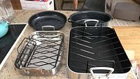 Photo showing three roasting pans: a large black enamel pan, a medium stainless steel La Creuset roaster, and another stainless steel pan with a black interior and roasting rack.