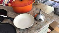 Orange Le Creuset Dutch oven with lid, white frying pan with black handle, black round perforated bakeware pan, and stainless steel funnel with orange handle on countertop.
