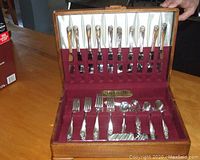 Cutlery set displayed in wooden chest with multiple slots holding knives, forks, and spoons showing daffodil pattern on handles and burgundy fabric interior.