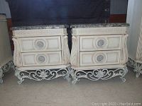Two cream-colored Pulaski bedside tables side by side showing marble-look tops, two drawers, ornamental drawer pulls, and wrought iron legs with scrollwork