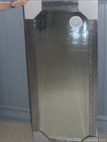 Full view of a rectangular mirror standing vertically, showing the black mosaic plastic frame and white corner guards.