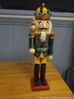 Front view of the 25-inch tall wooden nutcracker figurine with colorful crown, staff, and painted attire on a 5x5 inch red base.
